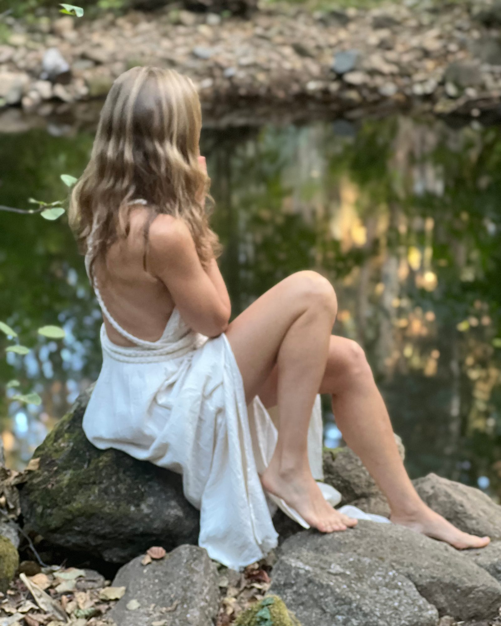 Miriam Ysa Calista von hinten, in einem weißen Kleid auf einem Felsen am Wasser sitzend